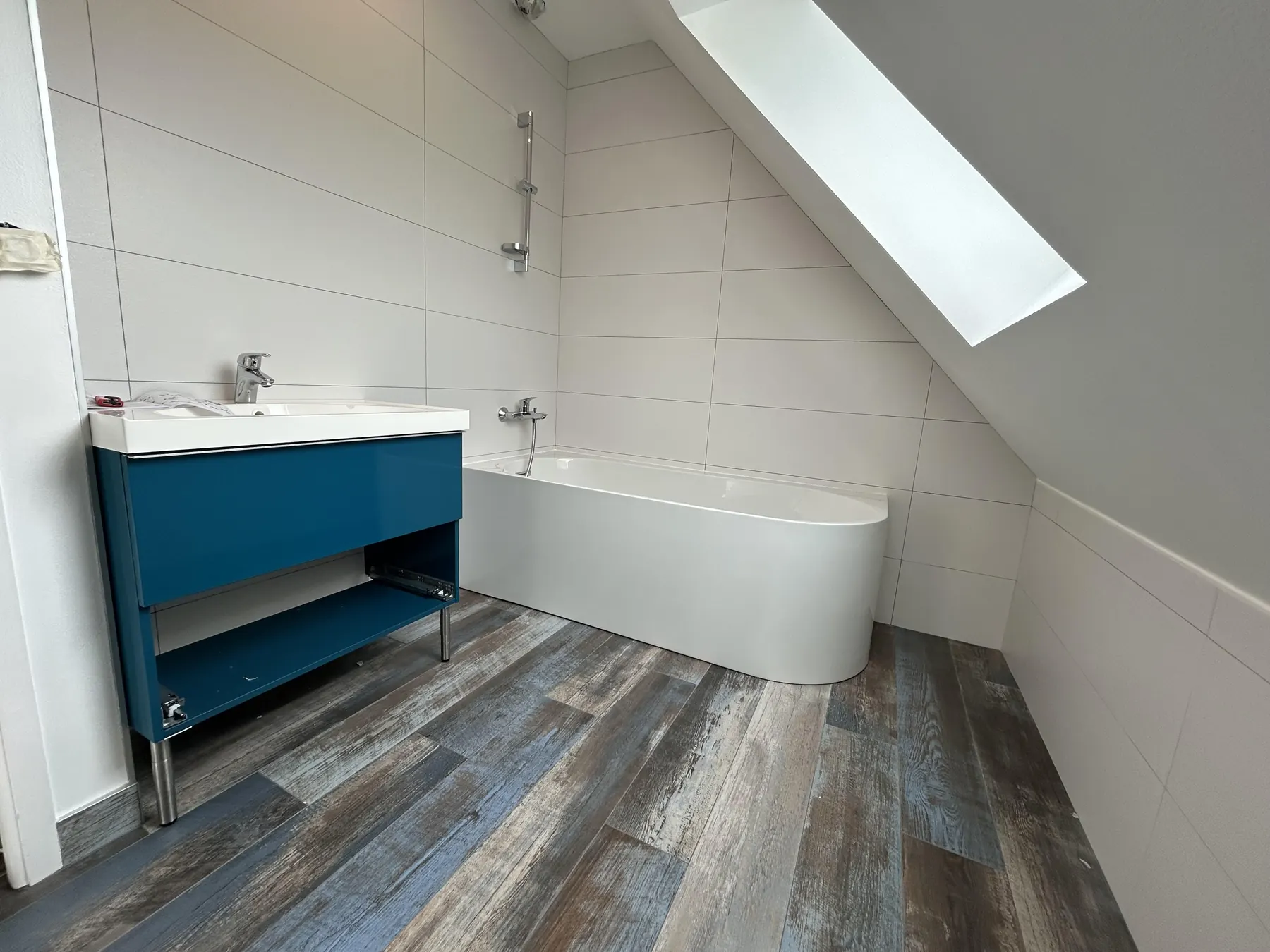 Salle de bain sous combles avec baignoire, meuble vasque bleu et sol effet bois
